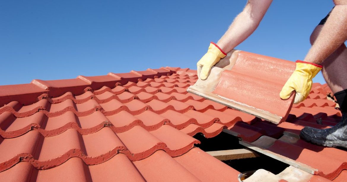 Roof repair, worker with yellow gloves replacing red tiles or shingles on house with blue sky as background